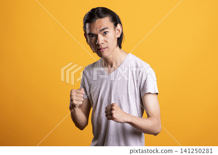 Frustrated asian man isolated over yellow background, shows fierce emotion and maintains fighting stance. Serious guy poses with clenched fists, looking ahead with fearless expression, ready to fight. Frustrated asian man isolated over yellow background, shows fierce emotion and maintains fighting stance. Serious guy poses with clenched fists, looking ahead with fearless expression, ready to fight. 141250281
