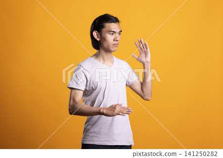 Young male adult demonstrating controlled and steady posture, appearing concentrated and ready in studio. Asian man stands in martial arts pose with one hand extended and other held close for balance. Young male adult demonstrating controlled and steady posture, appearing concentrated and ready in studio. Asian man stands in martial arts pose with one hand extended and other held close for balance. 141250282