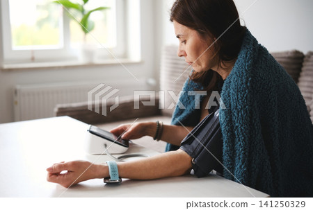Woman Sitting On A Sofa In A Blanket Turning On A Digital Blood Pressure Monitor And Feeling Unwell Woman Sitting On A Sofa In A Blanket Turning On A Digital Blood Pressure Monitor And Feeling Unwell 141250329