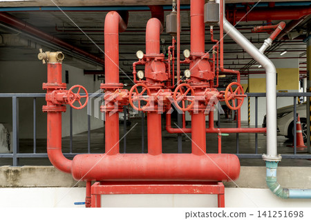 Red industrial fire protection pipes featuring valves and pressure gauges in a parking garage. Professional fire sprinkler pipeline installation for building safety and emergency water response. Red industrial fire protection pipes featuring valves and pressure gauges in a parking garage. Professional fire sprinkler pipeline installation for building safety and emergency water response. 141251698