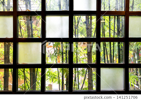 Traditional window with a dark wooden grid pattern looking out onto a lush green bamboo forest. Some panes are frosted, creating a serene and zen-like Japanese garden atmosphere inside the room. Traditional window with a dark wooden grid pattern looking out onto a lush green bamboo forest. Some panes are frosted, creating a serene and zen-like Japanese garden atmosphere inside the room. 141251706