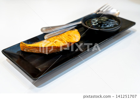 Grilled salmon fillet served on a sleek black rectangular plate with a side of savory soy sauce. Includes a silver fork and spoon, presented on a clean white background. Healthy seafood meal concept. Grilled salmon fillet served on a sleek black rectangular plate with a side of savory soy sauce. Includes a silver fork and spoon, presented on a clean white background. Healthy seafood meal concept. 141251707