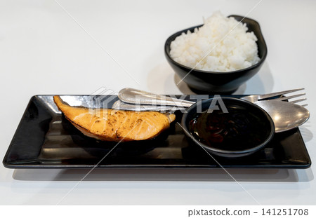 Grilled salmon teriyaki fillet served on a black rectangular plate with a bowl of steamed white rice and soy sauce. Traditional Japanese cuisine meal set isolated on a pure clean white background. Grilled salmon teriyaki fillet served on a black rectangular plate with a bowl of steamed white rice and soy sauce. Traditional Japanese cuisine meal set isolated on a pure clean white background. 141251708