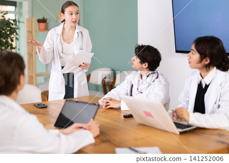 Confident young woman doctor standing near table where sitting medical interns talking and discussing ideas in medical office Confident young woman doctor standing near table where sitting medical interns talking and discussing ideas in medical office 141252006