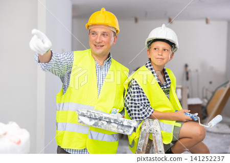 Portrait of father and son in yellow vest and protective helmet, who paint walls of room with paint roller Portrait of father and son in yellow vest and protective helmet, who paint walls of room with paint roller 141252237