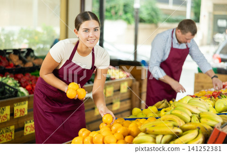 Happy girl seller working in supermarket and lays out fresh oranges on counter Happy girl seller working in supermarket and lays out fresh oranges on counter 141252381