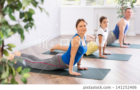 Happy middle-aged woman practicing cobra pose of yoga with her family Happy middle-aged woman practicing cobra pose of yoga with her family 141252406