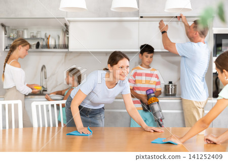 Friendly large family cleans room together. Mom and daughter are dusting table. Son is vacuuming oom. Father together with daughters wipe dust 141252409