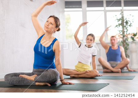 Happy middle-aged woman practicing half lotus pose of yoga with her family Happy middle-aged woman practicing half lotus pose of yoga with her family 141252454