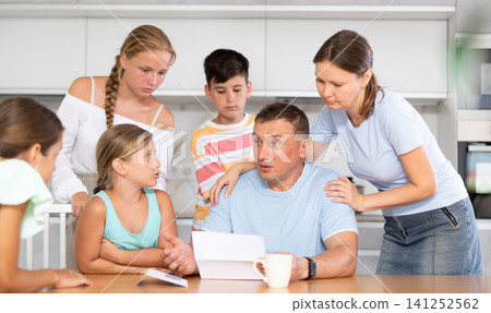 Frustrated man sits in kitchen next to family and reads unpleasant news in letter. Frustrated man sits in kitchen next to family and reads unpleasant news in letter. 141252562