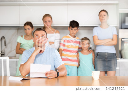 Upset father reads and looks through financial documents. Sad wife with children in background Upset father reads and looks through financial documents. Sad wife with children in background 141252621