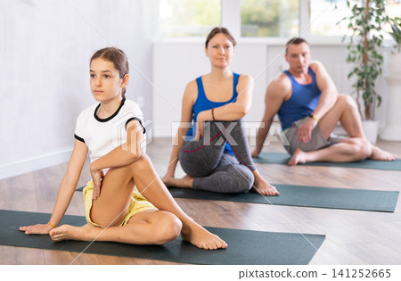 Pleasant preteen girl practicing half lord of the fishes pose of yoga with her family Pleasant preteen girl practicing half lord of the fishes pose of yoga with her family 141252665