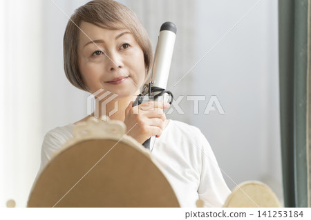 Senior woman styling her hair with a curling iron. Senior woman styling her hair with a curling iron. 141253184