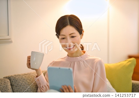 一位四十多歲的女性晚上在房間裡放鬆，看著平板電腦。 141253717