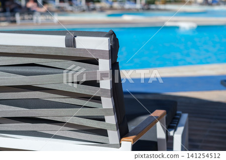 Modern outdoor lounge chair with gray woven straps placed beside a clear swimming pool, showcasing a serene and inviting leisure setting Modern outdoor lounge chair with gray woven straps placed beside a clear swimming pool, showcasing a serene and inviting leisure setting 141254512