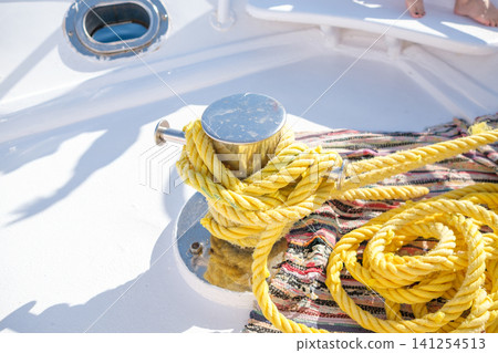 Yellow nautical rope is neatly coiled around a metal cleat on a boat deck, with a textured fabric and sunlight illuminating the scene Yellow nautical rope is neatly coiled around a metal cleat on a boat deck, with a textured fabric and sunlight illuminating the scene 141254513