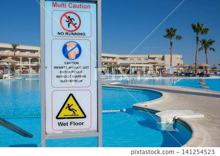 Multi-language caution sign displays safety warnings near a swimming pool, featuring a tropical resort and palm trees in the background 141254523