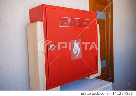 Red fire safety cabinet is mounted on a wall beside a wooden door, featuring fire safety symbols and instructions for emergency situations Red fire safety cabinet is mounted on a wall beside a wooden door, featuring fire safety symbols and instructions for emergency situations 141254536