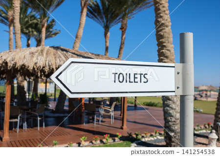 Sign pointing to toilets with clear lettering, surrounded by palm trees and a thatched roof structure in a recreational outdoor setting Sign pointing to toilets with clear lettering, surrounded by palm trees and a thatched roof structure in a recreational outdoor setting 141254538