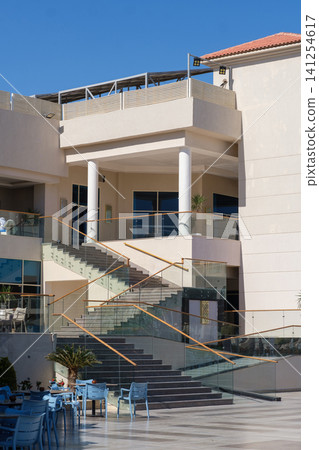 Modern commercial building architecture showcases a staircase with glass railings and outdoor seating area under bright sunlight Modern commercial building architecture showcases a staircase with glass railings and outdoor seating area under bright sunlight 141254617