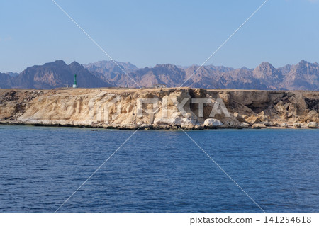 Rocky coastal area features a prominent cliff with mountains in the background and tranquil blue water in the foreground under a clear sky 141254618