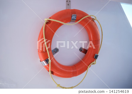 Bright orange life buoy is securely mounted on a white wall, equipped with metal brackets and a yellow rope for marine safety 141254619
