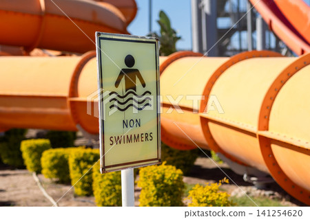Non-swimmers warning sign displayed beside orange water slides at a water park, surrounded by green shrubs and under a clear blue sky Non-swimmers warning sign displayed beside orange water slides at a water park, surrounded by green shrubs and under a clear blue sky 141254620