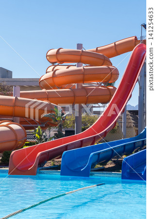 Vibrant water slides in a swimming pool area with clear blue sky, showcasing orange, red, and blue slides designed for recreational enjoyment Vibrant water slides in a swimming pool area with clear blue sky, showcasing orange, red, and blue slides designed for recreational enjoyment 141254633