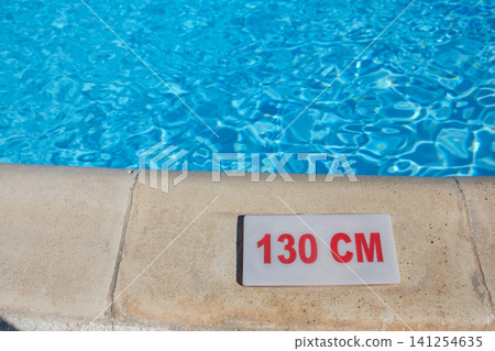 Swimming pool edge features a depth marker reading 130 cm, with vibrant blue water reflecting sunlight in the background Swimming pool edge features a depth marker reading 130 cm, with vibrant blue water reflecting sunlight in the background 141254635
