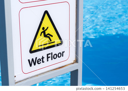 Caution sign warns of wet floor near a swimming pool, featuring a yellow triangle and a figure slipping, with blue water in the background Caution sign warns of wet floor near a swimming pool, featuring a yellow triangle and a figure slipping, with blue water in the background 141254653