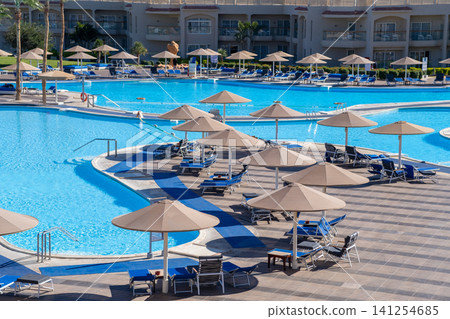 Resort swimming pool features sun loungers and umbrellas surrounding clear blue water, with modern architecture visible in the background Resort swimming pool features sun loungers and umbrellas surrounding clear blue water, with modern architecture visible in the background 141254685