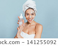 Woman with towel wrapped around head smiles while holding skincare product 141255092
