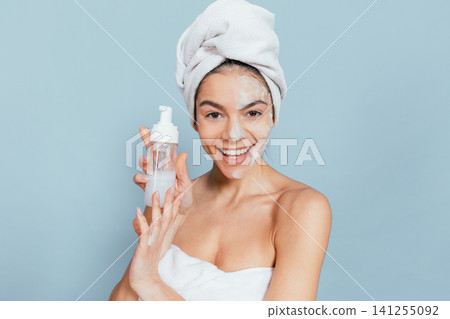 Woman with towel wrapped around head smiles while holding skincare product Woman with towel wrapped around head smiles while holding skincare product 141255092