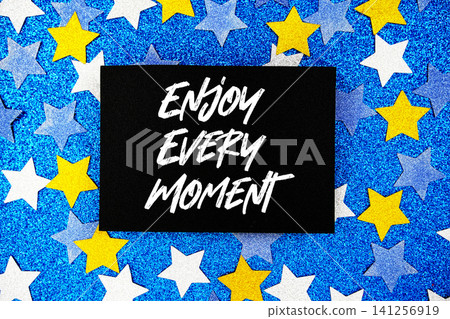 Enjoy Every Moment text written on paper card with star glitter on blue background Enjoy Every Moment text written on paper card with star glitter on blue background 141256919