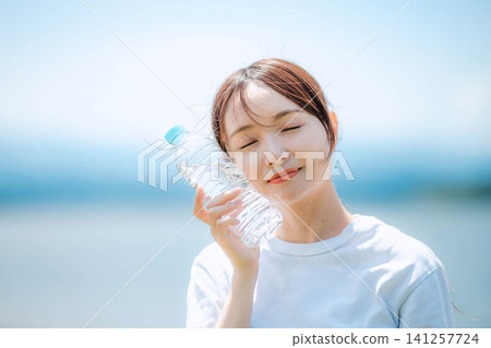 A woman cools herself by holding a plastic bottle against her cheek in a horizontal position. A woman cools herself by holding a plastic bottle against her cheek in a horizontal position. 141257724