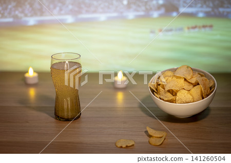 A glass of beer and potato chips snack on wooden table during soccer match, highlighting comforting food for big football events. A glass of beer and potato chips snack on wooden table during soccer match, highlighting comforting food for big football events. 141260504
