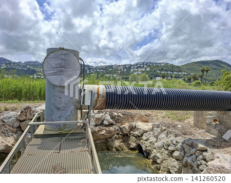 A water pumping station sits on a service walkway beside a corrugated pipe. It overlooks the green hills of the Pego Oliva ecological rice farming region under a cloudy sky. A water pumping station sits on a service walkway beside a corrugated pipe. It overlooks the green hills of the Pego Oliva ecological rice farming region under a cloudy sky. 141260520