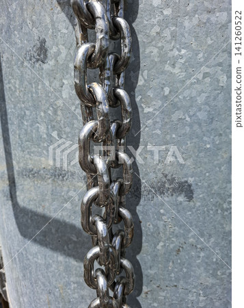 A heavy duty metallic steel chain hangs against a weathered galvanized metal background. It shows strength and security typical of industrial settings. 141260522