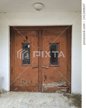A worn orange metal double door with peeling paint. The door has small rectangular windows and stands against a decayed wall of an abandoned hospital. 141260647