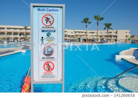 Multi-caution safety sign displayed near a swimming pool, featuring rules for safety, with resort buildings and palm trees visible in the background Multi-caution safety sign displayed near a swimming pool, featuring rules for safety, with resort buildings and palm trees visible in the background 141260658