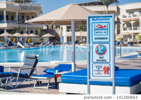 Swimming pool area features a multifunction sign with depth and safety rules, lounge chairs, and umbrellas under a clear blue sky Swimming pool area features a multifunction sign with depth and safety rules, lounge chairs, and umbrellas under a clear blue sky 141260663
