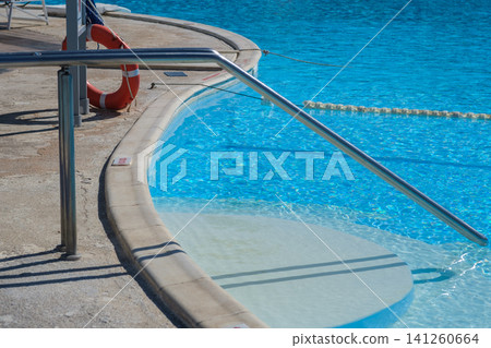 Swimming pool edge features a stainless steel railing and a lifebuoy, with clear blue water reflecting sunlight in an outdoor setting 141260664