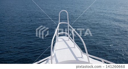 Bow view of a white yacht on tranquil blue ocean waters, highlighting the boat's railing and deck design against a clear sky backdrop Bow view of a white yacht on tranquil blue ocean waters, highlighting the boat's railing and deck design against a clear sky backdrop 141260668