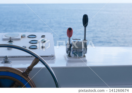 Boat control panel displays throttle lever, steering wheel, and gauges with a view of calm ocean waters in the background Boat control panel displays throttle lever, steering wheel, and gauges with a view of calm ocean waters in the background 141260674