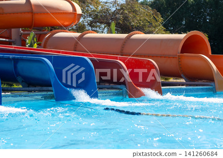 Colorful water slides are positioned beside a swimming pool with clear blue water and greenery visible in the background under bright sunlight Colorful water slides are positioned beside a swimming pool with clear blue water and greenery visible in the background under bright sunlight 141260684