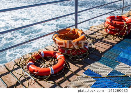 Lifebuoys are placed on a wooden deck by the ocean, with ropes coiled around them and waves visible in the background Lifebuoys are placed on a wooden deck by the ocean, with ropes coiled around them and waves visible in the background 141260685