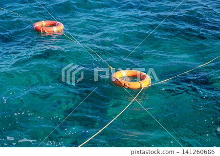 Two orange life rings are visible in clear blue water, connected by a rope, representing safety equipment in aquatic settings Two orange life rings are visible in clear blue water, connected by a rope, representing safety equipment in aquatic settings 141260686