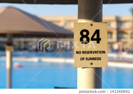 Sign with the number 84 stating no reservation of sunbeds, positioned near a swimming pool with umbrellas and water in the background Sign with the number 84 stating no reservation of sunbeds, positioned near a swimming pool with umbrellas and water in the background 141260692