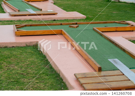 Miniature golf course design with green turf and wooden barriers, set on a manicured lawn in an outdoor recreational area Miniature golf course design with green turf and wooden barriers, set on a manicured lawn in an outdoor recreational area 141260708