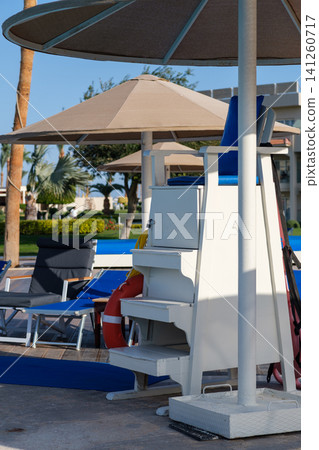 Lifeguard chair equipped with safety gear alongside lounge chairs and umbrellas near a swimming pool at a resort during daytime Lifeguard chair equipped with safety gear alongside lounge chairs and umbrellas near a swimming pool at a resort during daytime 141260717